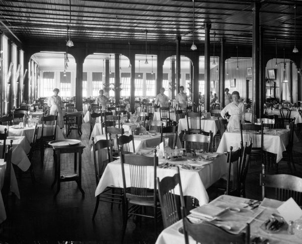 Interior of a dining room at Forest Park Hotel, purchased by the International Ladies' Garment Workers Union in the early 1900's. Women stand among tables and chairs set for a meal.