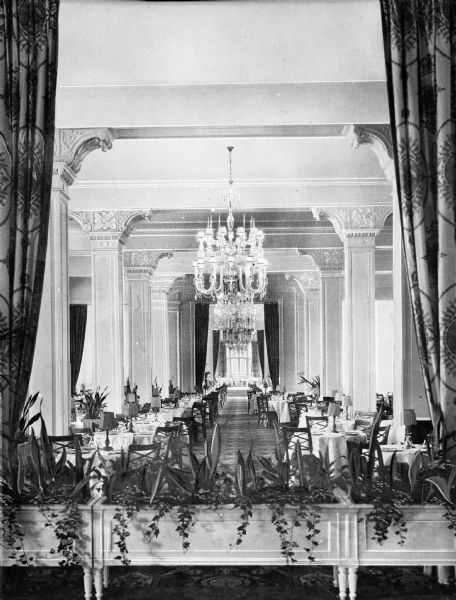 Interior of a dining room at Greenbrier Hotel, built in 1913. View from behind drapes and plants in plant stands toward tables and chairs which fill the room. Beyond rows of columns is a window at the far end of the room.