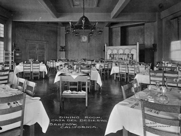 Interior of a dining room at Casa Del Desierto, opened in 1911 by Fred Harvey. The room features tables, chairs, a chandelier, and decoration in a Spanish motif. Caption reads: "Dining Room, Casa Del Desierto, Barstow, California."