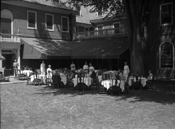 Exterior of Exeter Inn with guests dining outdoors under trees. Built in 1932, the hotel's Georgian facade features a doorway and an awning under which tables are set for a meal.