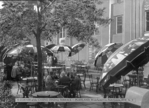 A corner of the Casino Dining Terrace at Playland, part of the Westchester County Park System. Built in 1928 by Frank Darling, the cafe at the terrace holds groups of people sitting at tables under umbrellas. Caption reads: "A Corner of the Casino Dining Terrace - Playland, Westchester Co. Park System, Rye, N.Y."
