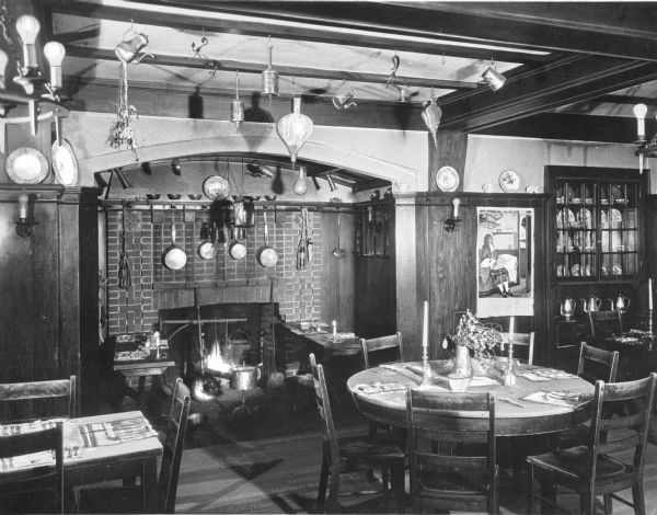 Interior of an Early American dining area at Wellesley Inn, opened in 1910 and designed by architect Donald Ross. The room features a fireplace surrounded by tables and chairs set for a meal. Pots hang from hooks over the fireplace, tea kettles, and fireplace bellows, and other utensils hang from the ceiling.