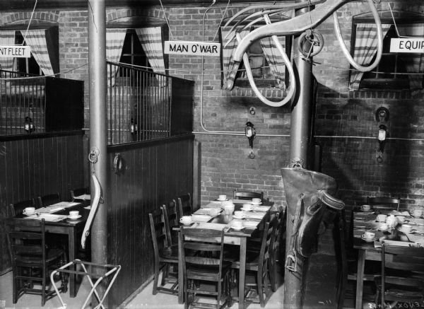 Interior of a restaurant decorated with western-like decor. Tables and chairs are surrounded by stalls, saddles, and harnesses, with draped windows in the brick walls. A sign above one section is labeled, "Man O'War."