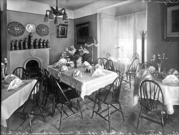 Interior of a restaurant dining room. Decorative steins are placed on the mantle of a fireplace, and tables and chairs are arranged around the room.
