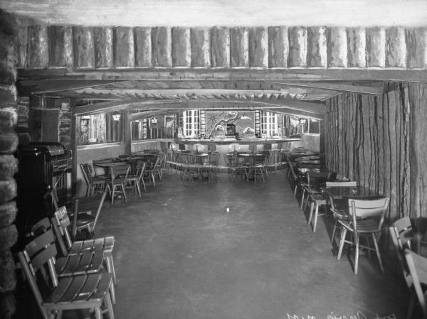 Interior of a bar in a rustic inn. Chairs are arranged along the walls and against a bar in the background and a jukebox stands along the wall at left.