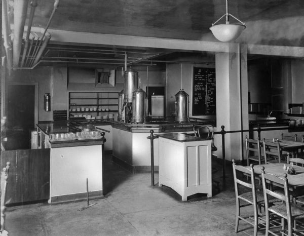 Interior of the YWCA Cafeteria, built in 1918. Kitchen equipment can be seen on counter tops and tables, and chairs stand to the right. On the back wall, a sign lists the menu.