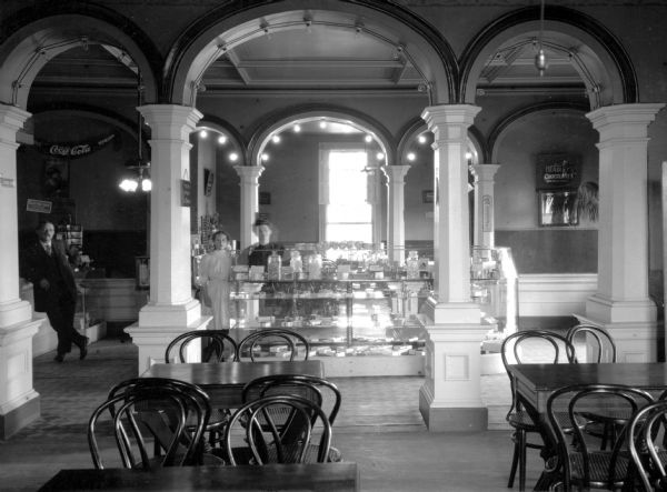 Interior of a restaurant at a mansion house. A man at the left leans against a counter, and two women stand behind a shelf filled with candy.  Tables and chairs fill the room and signs advertising Coca-Cola and Hadley's Chocolates hang in the background.