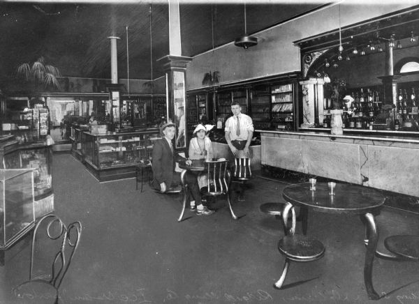 Two men and a woman sit and stand around a table inside the Ridgell Drug Company and ice cream parlor. Several display cases are arranged behind them and the ice cream parlor counter stands to the right.