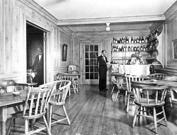 Interior of the bar at the Terry Tavern. Waiters stand near the bar and a man looks in from the room on the left.  Tables and chairs fill the bar room.