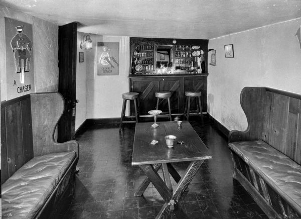 A bar counter and stools stand inside a barroom and several posters adorn the walls.  In the foreground, two cushioned benches are placed on either side of a wooden table.