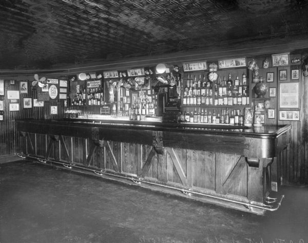 Interior of a rustic barroom with tin ceiling. Bottles of alcohol are arranged on shelves behind the wooden bar and framed artwork hangs on the walls.