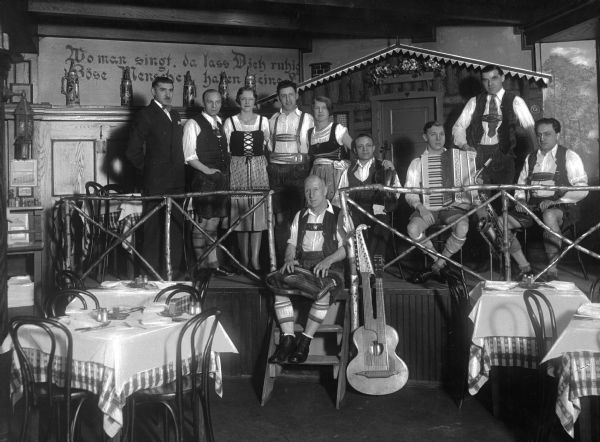 Interior of the Brau Haus, a German-American Rathskeller.  A German band wearing costumes stands on the small stage at the restaurant, some holding musical instruments.