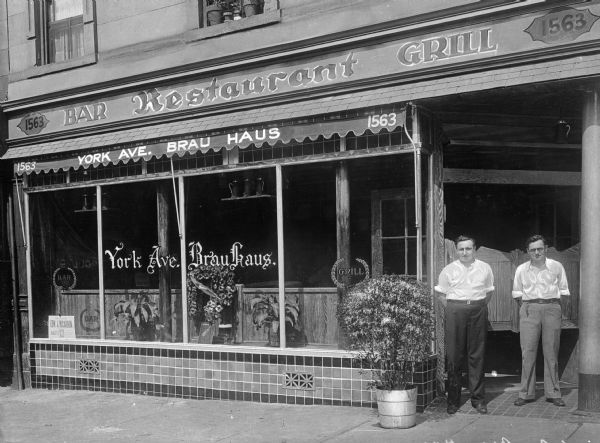 Two men stand in a doorway near a potted plant outside the Brau Haus, a German-American Rathskeller.  The lettering painted on the window reads, "1563 York Ave. Brau Haus, Bar, Restaurant, Grill."