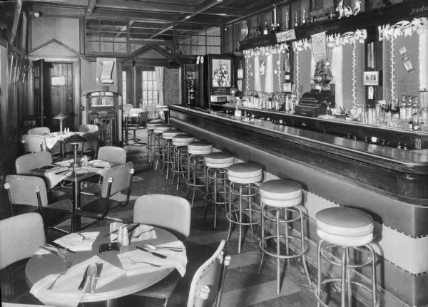 Interior of Mike Mazie's Bar. A counter and stools can be seen to the right of a dining area arranged with tables and chairs. In the background, a jukebox can be seen near a door.