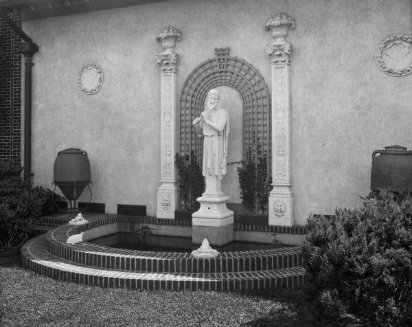 A sculpture of a man playing a flute adorns a fountain against the exterior wall of the Biltmore Club.  Engaged columns and an arch can be seen behind the sculpture.  The Biltmore Club was established in 1922.