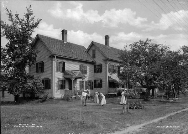 Men and woman stand outside a mountain view house while playing croquet on the front lawn.  Published by Fred W. Brown.