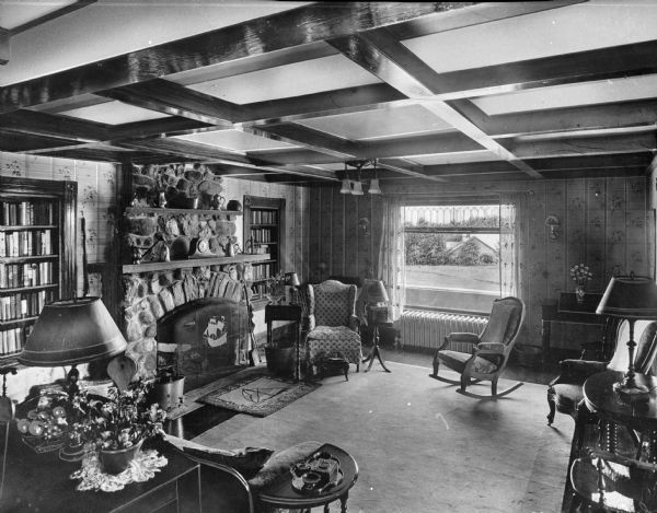 Interior of the Spalding Inn, furnished with chairs and end tables arranged around a fireplace with a stone mantle.  Recessed bookshelves stand on either side of the fireplace and, through the window on the far wall, a residential building can be seen.