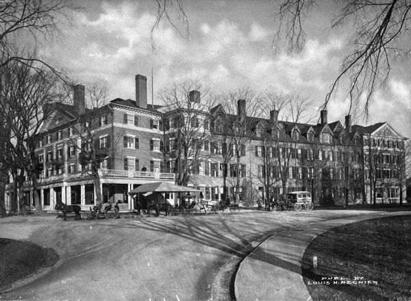 Exterior of the Curtis Hotel, a Victorian era building expanded in 1883.  Horse-drawn carriages can be seen parked underneath an awning, and a man stands on the balcony above.  Published by Louis H. Regnier.