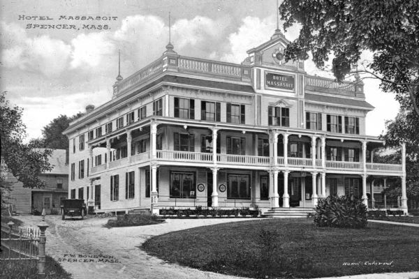 Exterior of Hotel Massasoit, built in 1887.  The wooden building features a long porch and balcony.  To the left of the hotel, an automobile is parked.