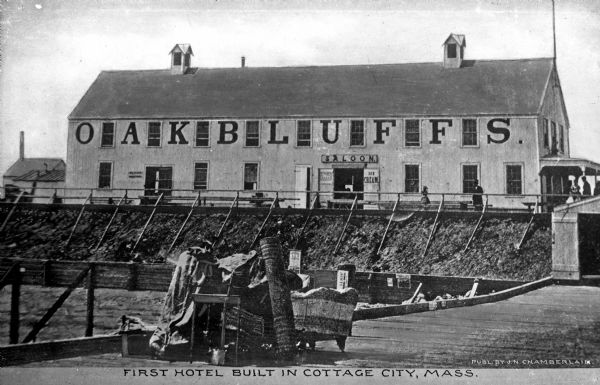 View of Oak Bluffs Hotel from a waterfront boardwalk.  Pedestrians walk along the boardwalk in front of the hotel and furniture and other luggage stands in a pile on the dock in the foreground.  The signs near the hotel entrance read, "Saloon," "Ice Cream," and "... Beer."  Caption reads: "First Hotel Built in Cottage City."  Published by J.N. Chamberlain.