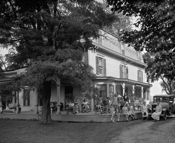 A group of girls and women sit on the porch of the Holiday House of the Girls' Friendly Society while others sit inside an automobile at right to pose for a group portrait.  The Girls' Friendly Society was founded in 1875.