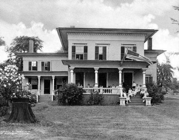 A group of adults and children sit on the porch of The Elms, a two-story house with an American flag flying from the porch roof.  Tree stumps are in the front yard and the house is surrounded by trees and flowering bushes.
