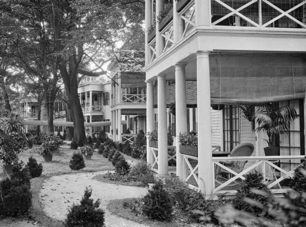 View of the rear porches and balconies at Hilltop Inn, built in 1910. Trees and other plants surround the porches where wicker chairs and potted plants are arranged.