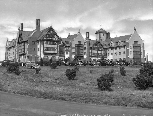 Guests walk toward the entrance of Montauk Manner and automobiles are parked in front of the building.  The English Tudor-style hotel opened in 1927.