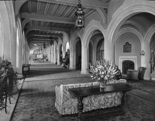 View down a long hall separated by arches and columns in the lobby of Montauk Manor.  The space is furnished with couches, chairs, and a fireplace and a table holds a floral arrangement in the foreground.  The English Tudor-style hotel opened in 1927.