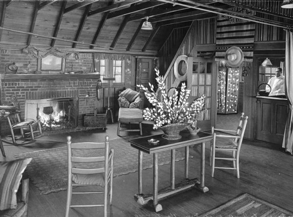 Chairs and tables are arranged around a fireplace in a lounge area at Rulueys Inn.  A man stands waiting at the reception desk on the right.