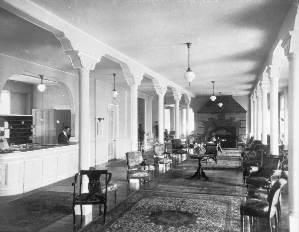 Interior of the lobby at Poggatticut Lodge, built in 1920.  The waiting area features decorative, upholstered chairs and a fireplace, and a man stands behind the reception desk on the left.