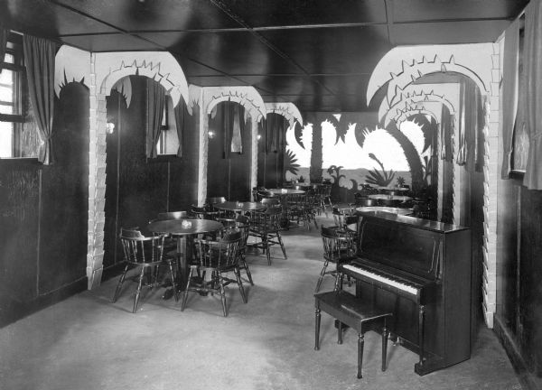 Interior of the Hawaiian Room at Hanson Hotel, one of the original structures built near Oquaga Lake.  The room is decorated with palm trees that protrude from the side walls and a Hawaiian scene made from cut paper is visible on the far wall.  A piano, chairs, and tables fill the room.