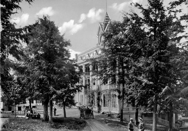 Men and women stand outside the Laurel House as horse-drawn carriages drive past the building along a dirt road. The structure was built in 1852 by Peter Schutt.
