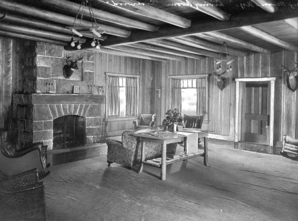 Chairs and a table are arranged around a fireplace inside the rustic living room at Covewood Lodge. The ceiling features exposed wooden beams and the walls are decorated with mounted deer heads. Covewood Lodge opened in 1924.