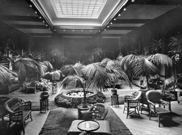 Wicker chairs, tables, and potted palms are arranged beneath a skylight in the lobby of Laurel-in-the-Pines Hotel. The hotel opened in 1891.