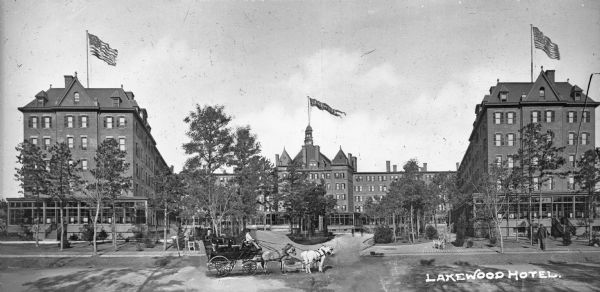 View across road toward the hotel. In the foreground is a horse-drawn carriage. The hotel opened in 1891 and the building features a central pavilion with two flanking buildings that extend toward the road. A flag reading "Lakewood" flies from the middle building and American flags fly from the adjacent rooftops. Caption reads: "Lakewood Hotel."