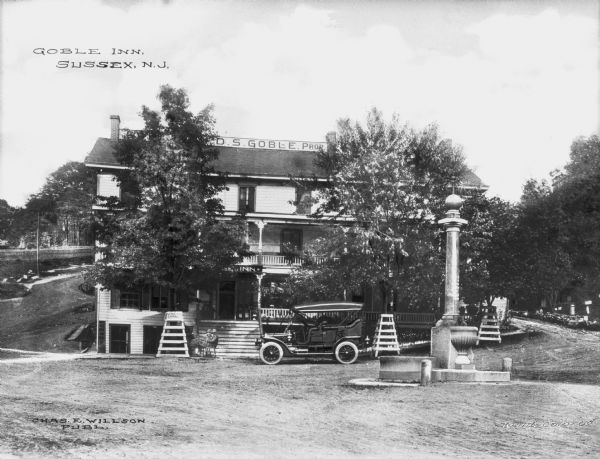 Exterior of the Goble Inn. The land, purchased in 1892, features a driveway and trees surrounding the inn. An automobile is parked near stairs that lead to the porch of the building. Signs read: "Goble Inn," and "D.S. Goble, Proprietor." In the foreground on the right is a fountain that was erected by the Martin Foundation of Sussex. Caption reads: "Goble Inn, Sussex, N.J."