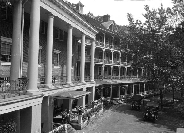 Elevated view looking along the front of the Bedford Springs Resort, built in 1806. Two automobiles are on the driveway near steps leading to the ground floor porch. Large columns are above the main entrance, and balconies are along each level of the resort.