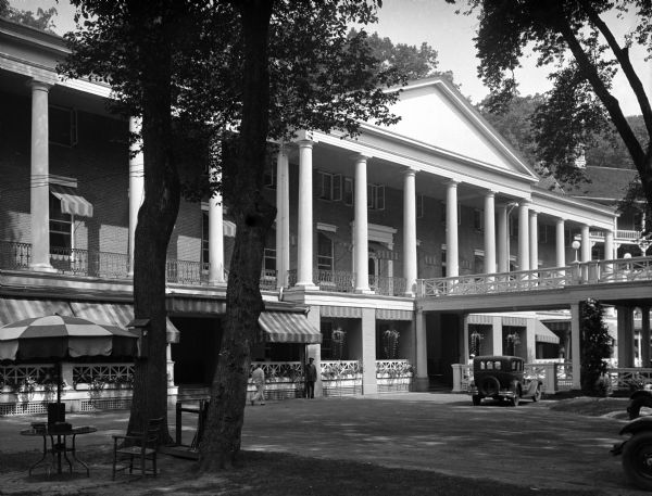 Exterior view of the Bedford Springs Resort, built in 1806. A car at the entrance is under a second-story walking bridge that leads to a large, balcony with columns and a pediment. Awnings are along the ground level porch. Chairs and a table with an umbrella are on the lawn under trees.