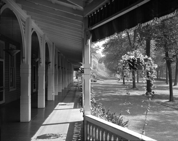 View of Bedford Springs Resort, built in 1806, taken from the veranda. A row of arched columns spans the length of the veranda between the front of the building on the left and a railing decorated with hanging potted plants in the center. A driveway on the right runs along the front of the hotel.