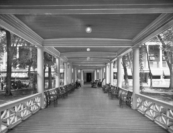 Exterior of Bedford Springs Resort, built in 1806. The view is from one end of a colonnade, with a band performing near a door that leads into the resort building. A man and child watch the musicians while a woman reads a newspaper. To the left, a group of men stands on the lawn near a parked automobile.