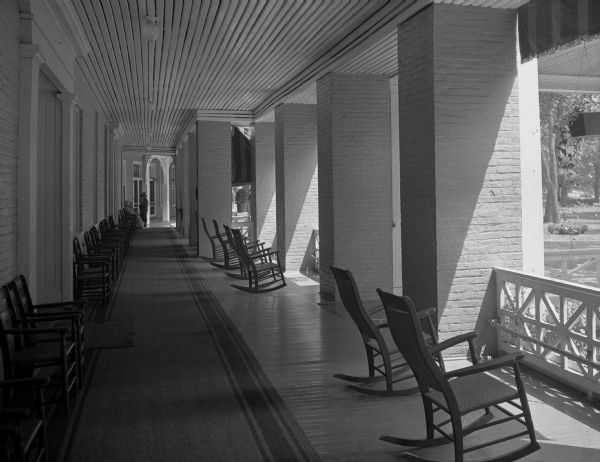 View of the lower veranda of Bedford Springs Resort, built in 1806. Straight backed chairs line the wall on the left, and rocking chairs are along a railing on the right. The railing is separated by large brick pillars. Three women are at the far end of the veranda near a door.