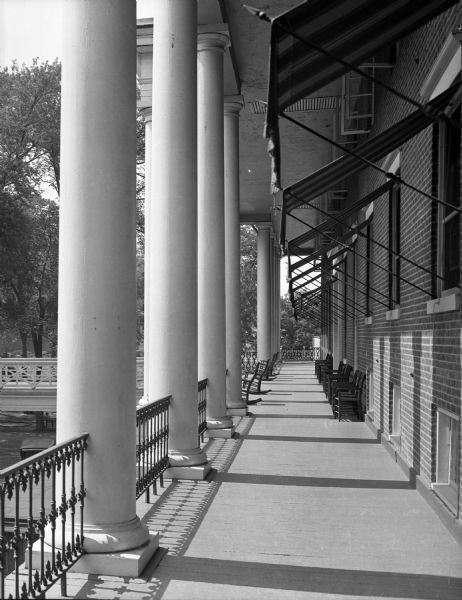 View along the front of the dining room veranda at Bedford Springs Resort, built in 1806. Straight backed chairs are arranged along the brick facade on the right underneath striped awnings above the windows.  Rocking chairs are along the railing in between the two-story columns. On the left, a walking bridge extends from the veranda over the driveway.