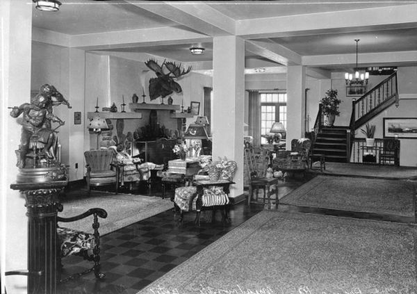 View across lobby area towards a woman sitting in an upholstered chair near a fireplace in the Meadowside Hotel. A mounted moose head hangs above the mantle and chairs and carpets are arranged throughout the rest of the lobby. A staircase is in the background.