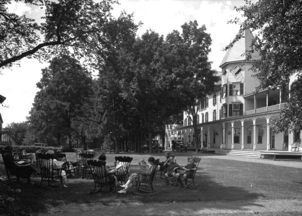 Adults and children read newspapers and books while sitting on chairs on the lawn outside Bluff House. The house is on the right, featuring a porch, balcony, and a octagonal central tower. The Bluff House was built in about 1876 by H.B. Wells.