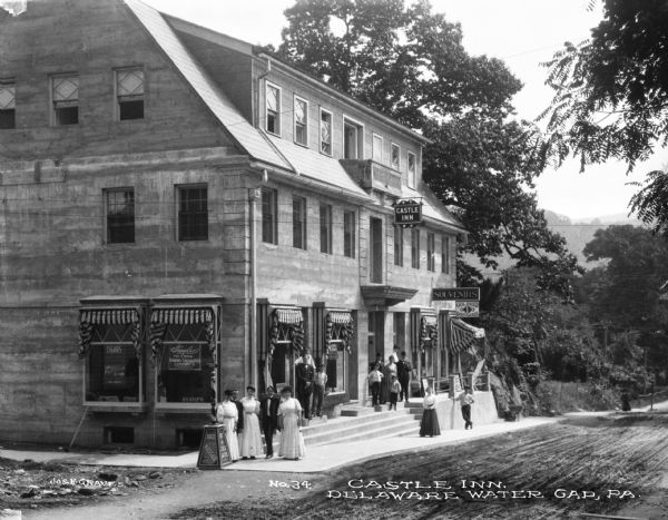 Slightly elevated view of children and adults standing on the sidewalk near the steps at the front of the Castle Inn. Signs over the windows adorned with awnings read: "Souvenirs" and "One Cent Postcards." A small balcony is just above the Castle Inn sign on the top floor. The Castle Inn opened in 1909.