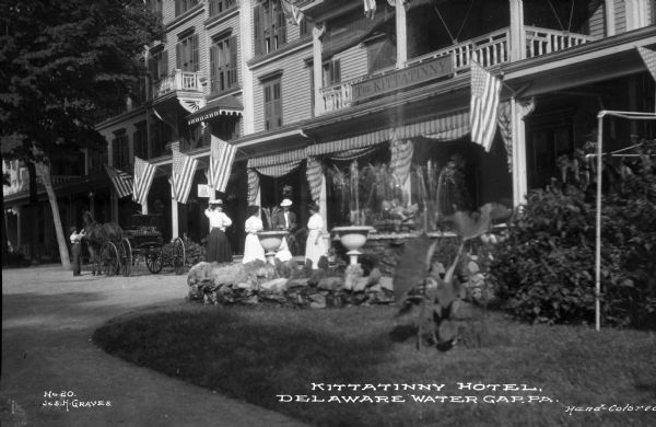 View from sidewalk toward men and women standing near the entrance to the Kittatinny Hotel while a boy tends to a horse and carriage to their left. The facade is adorned with American flags and a signboard that reads: "The Kittatinny." The hotel was built in 1872. Caption reads: "Kittatinny Hotel, Delaware Water Gap, PA."