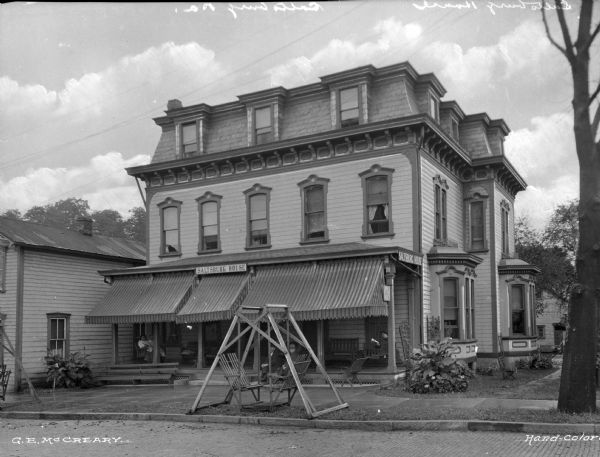 View across road toward men and women sitting on the porch of the three-story Saltsburg House hotel. Awnings are along the front of the porch, and a sign for the hotel is on the porch roof above the steps. A lawn swing is in front of the house along the curb.