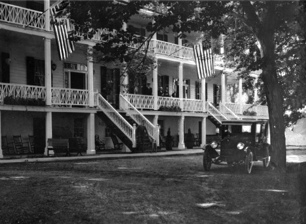 Exterior view from driveway toward the Mountain Springs Hotel, built in 1848. A man is at the base of a staircase leading to the porch of the building where four other men are standing. An automobile is parked near a tree on the lawn.