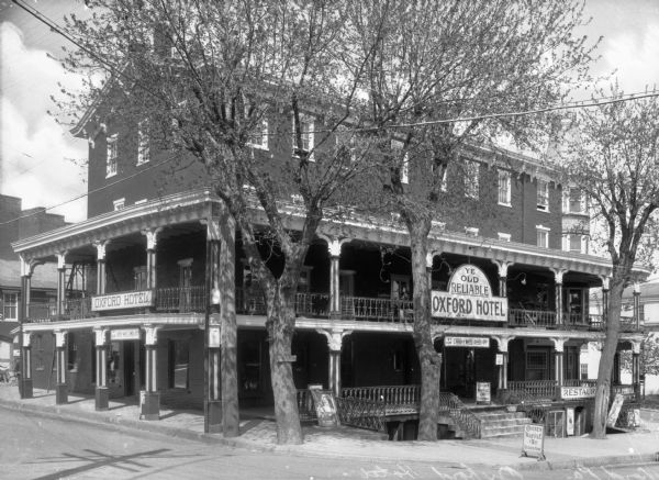 Exterior view from intersection toward the Oxford Hotel, built in 1858. Stairs on the right side lead to a porch, and a sign on the balcony reads, "Ye Old Reliable Oxford Hotel." Below is another sign that reads, "Try Our Chicken and Waffle Dinners $1.00." There is a sign on the left side of the hotel that reads, "Oxford Hotel."